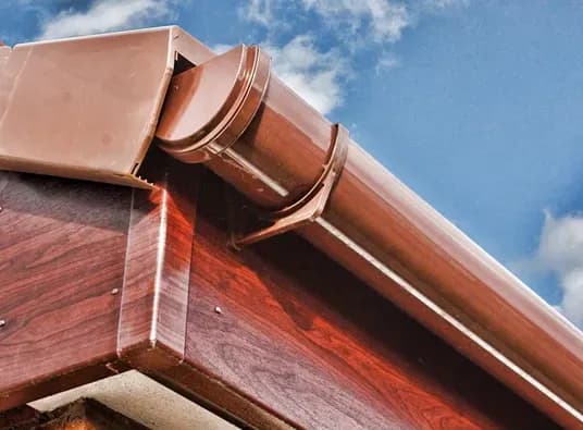 Roof corner with brown, wood-like cladding and matching rain gutter against a clear blue sky. The structure appears polished and sturdy.