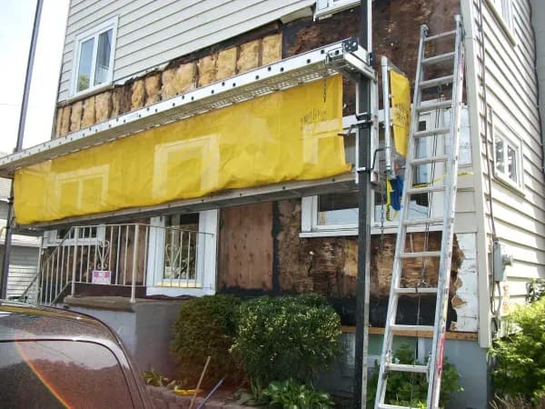 House undergoing renovation with siding removed, exposing dark, weathered wood. A ladder and scaffolding with a yellow tarp are set up in front.