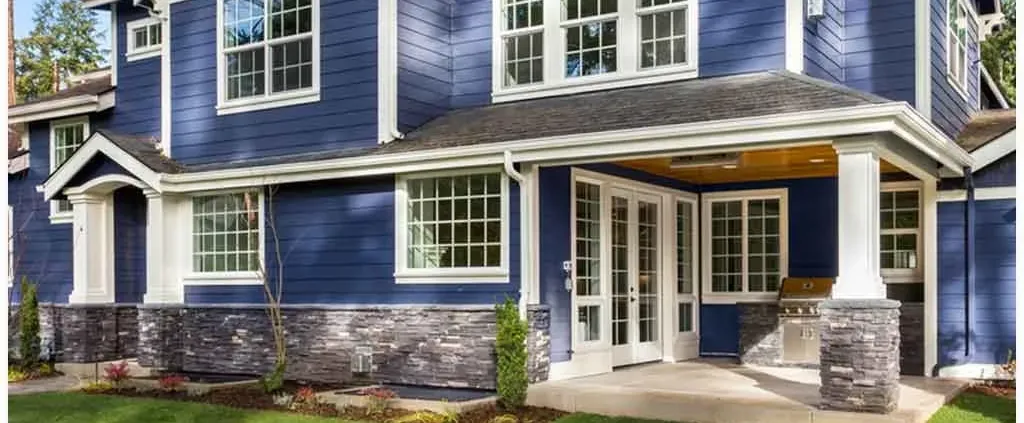 A modern two-story house with blue siding, large white-trimmed windows, and a stone accent base. The porch has white pillars and a built-in outdoor grill.