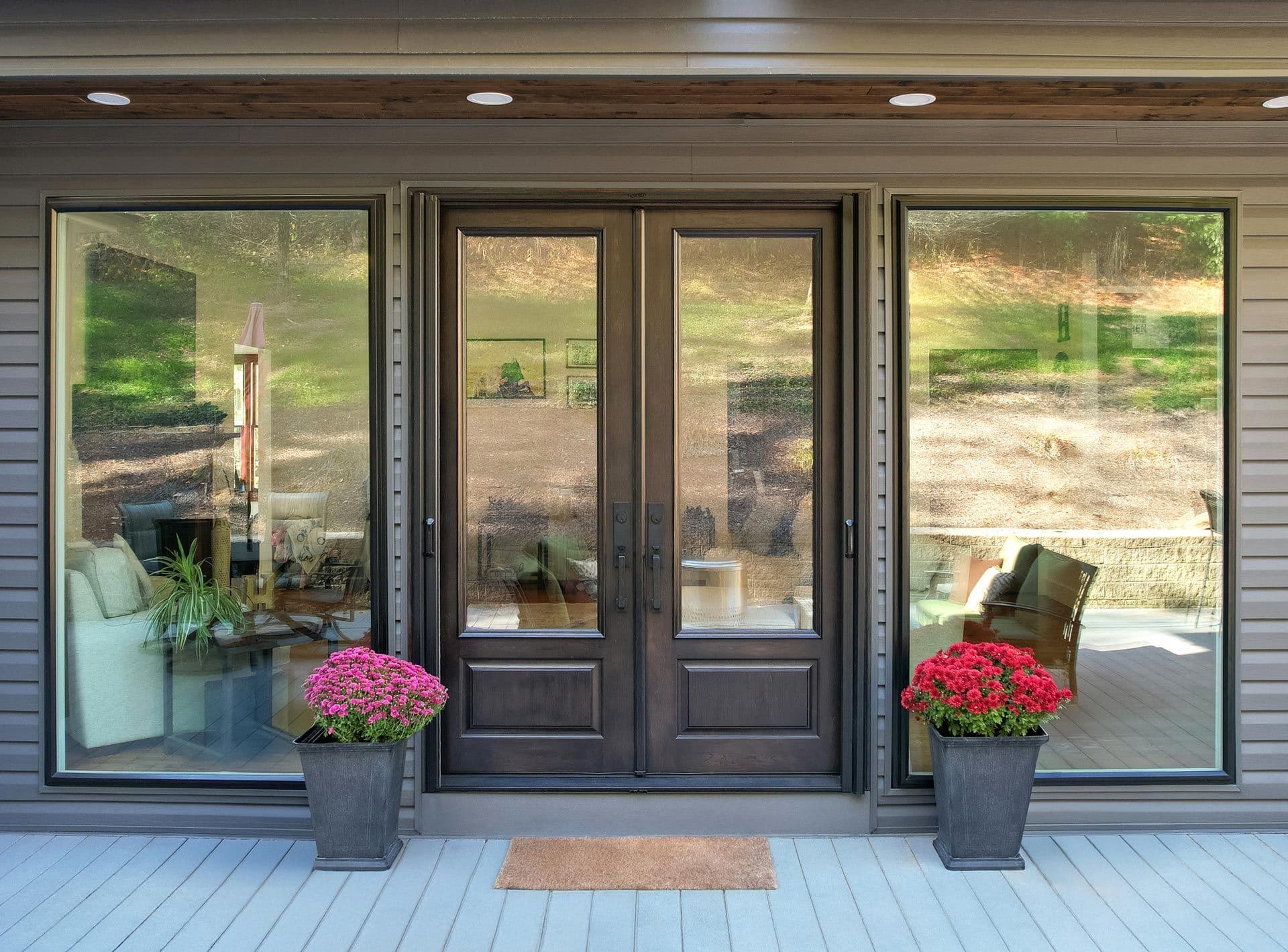 Large glass doors with dark frames open to a cozy interior, flanked by tall windows. Potted flowers on each side add color to the modern facade.