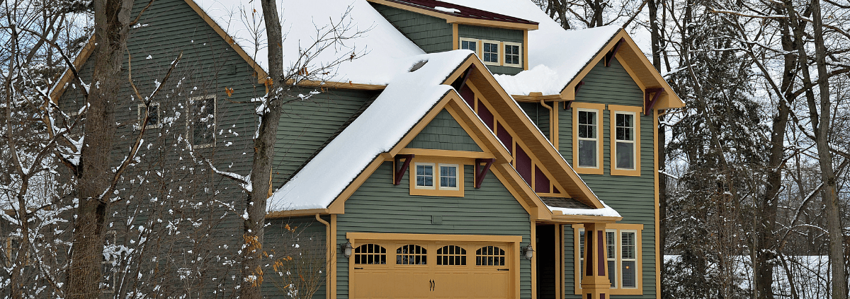 A charming green house with mustard trim stands amid snow-covered trees. The sloped roof and garage add character, creating a cozy, wintery atmosphere.