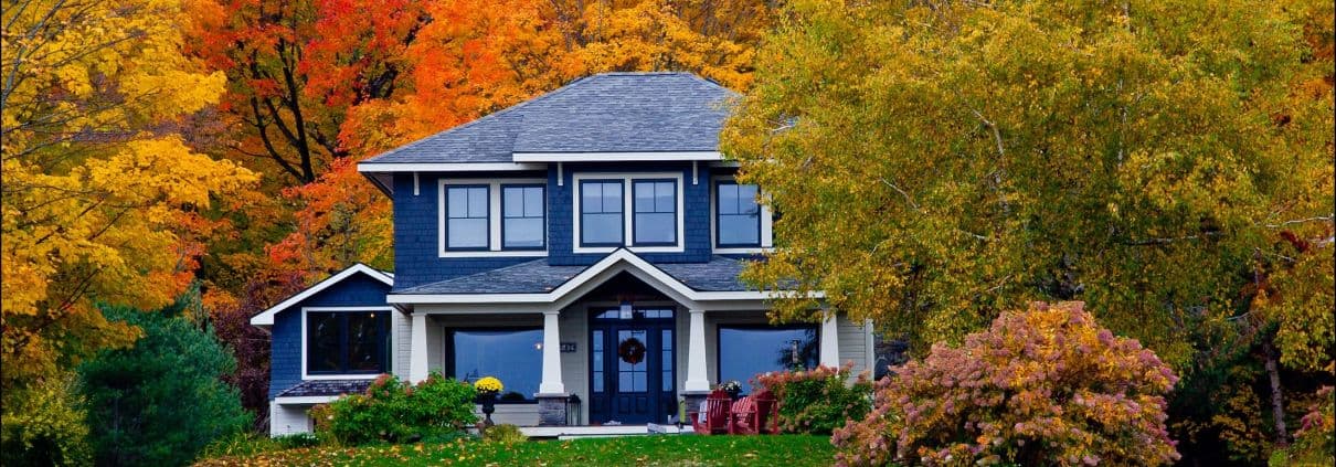 A charming two-story house with a dark blue exterior is surrounded by vibrant autumn foliage in shades of red, orange, and yellow, creating a cozy and inviting scene.