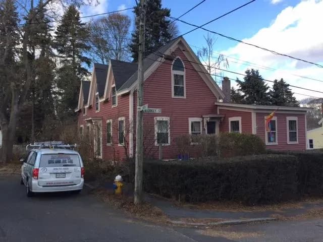 A red, two-story house with a gabled roof on a corner lot. A white van is parked nearby. A rainbow flag hangs near the entrance, and leafless trees surround the yard.