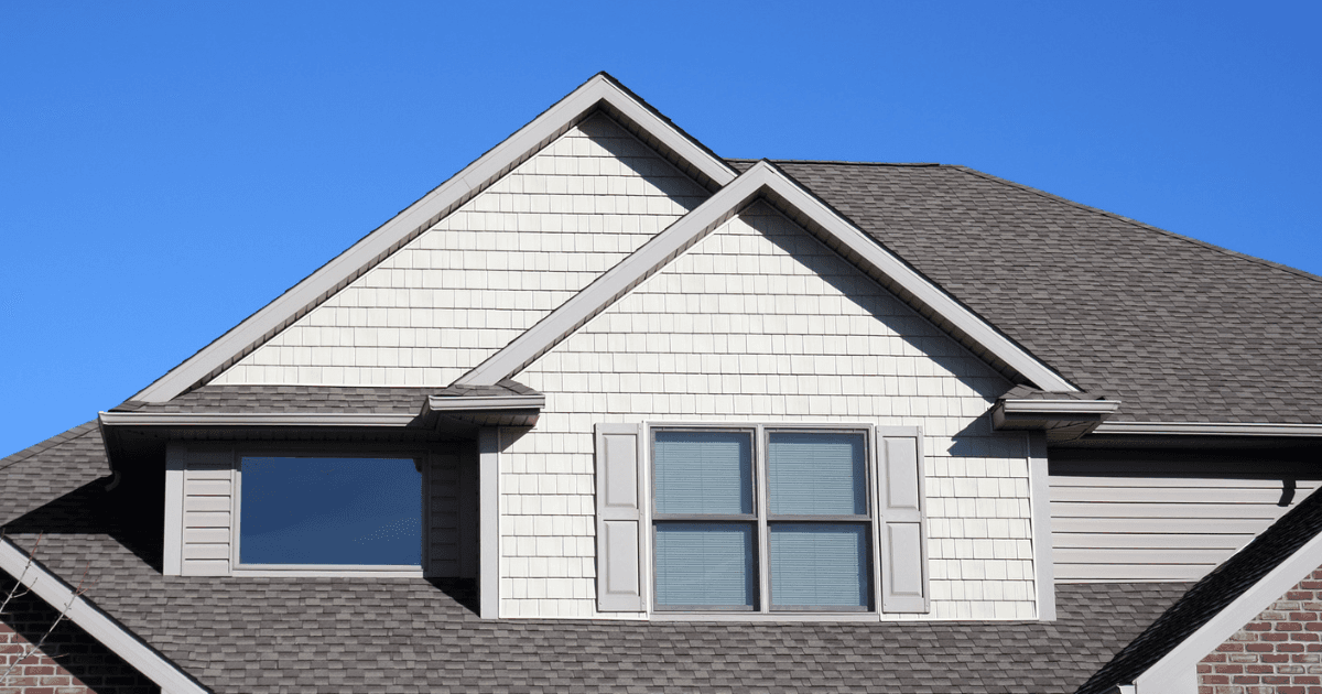 Roofline of a house with light beige siding and gray shingles under a clear blue sky. Features two windows and a section of brown brick wall.