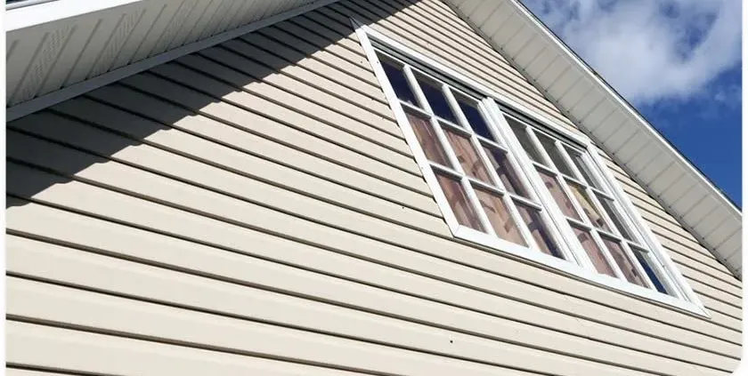The image shows a beige house facade with white trim and large grid windows. The roof edges frame the blue sky, evoking a bright, serene atmosphere.