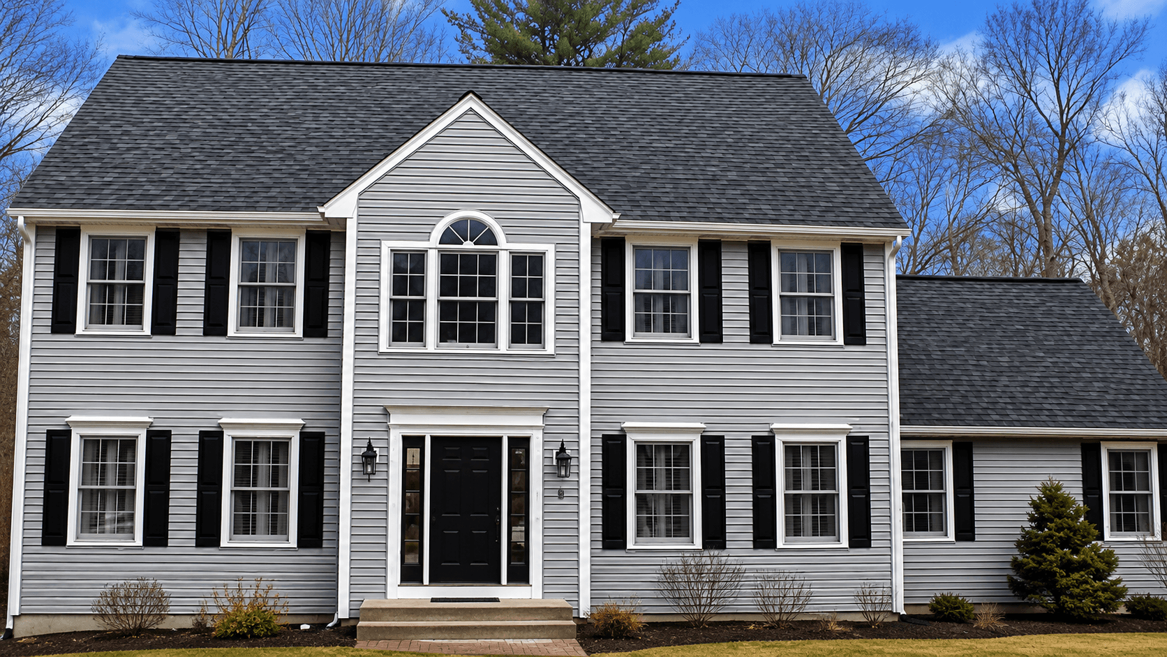 Two-story, gray house with black shutters, a peaked roof, and symmetrical windows. The front door is centered; trees and clouds are visible behind.