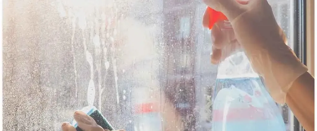 Hands wearing gloves clean a sunlit, dirty window using a spray bottle and sponge. The sunlight creates a warm, fresh atmosphere.