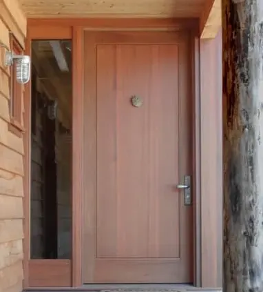 A wooden front door with a vertical glass panel on the left, metal handle, and small decorative emblem. Warm, inviting tones convey a rustic charm.