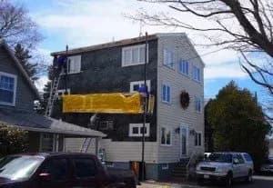 A three-story house with siding partially installed, revealing black underlay. A large yellow tarp hangs across the second floor. Two workers stand on ladders.