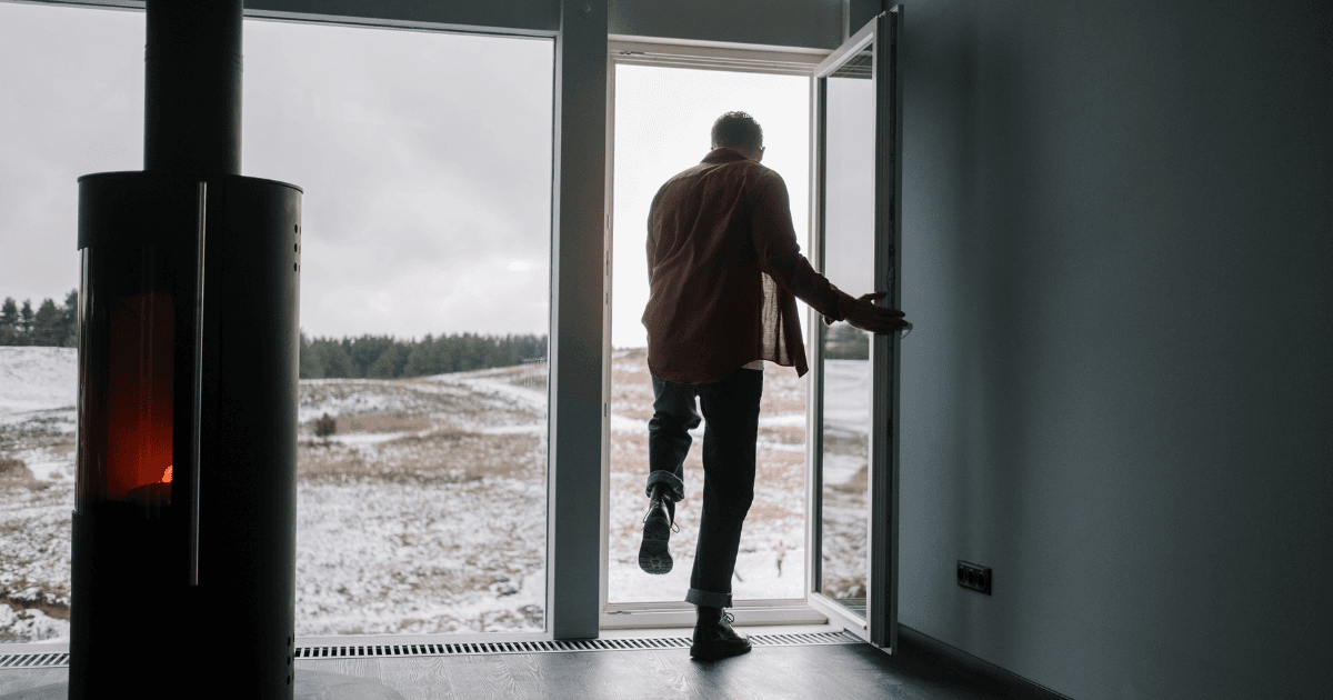 A person steps out a door of a cozy, dimly-lit room with a fireplace, into a snow-covered landscape. The scene conveys a sense of calm and transition.