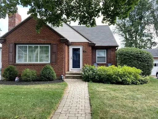 A charming brick house with a blue door, surrounded by lush green bushes and a tidy lawn. A stone path leads to the entrance, creating a welcoming feel.