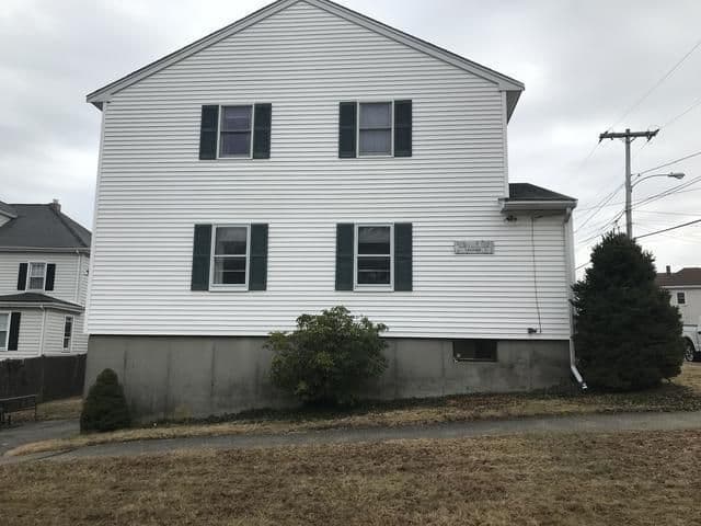 A two-story white house with dark shutters and a concrete foundation is shown on a cloudy day, surrounded by sparse grass and a few bushes.