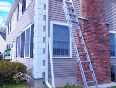 A wooden ladder leans against a two-story house, positioned near a red brick chimney. The house features beige siding, white trim, and shuttered windows.