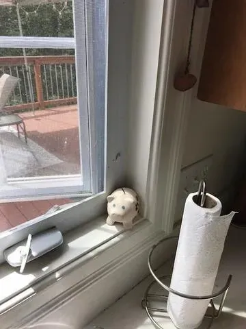 A small sheep figurine sits on a sunny window ledge beside a napkin holder and a phone charger. Outside, a deck with chairs is visible. Cozy kitchen scene.