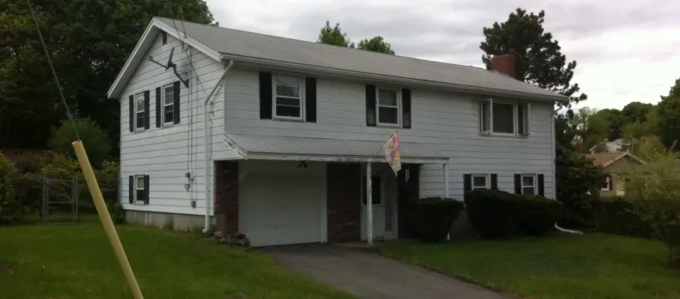 A two-story white suburban house with black shutters, a sloped roof, and a single garage. It's surrounded by green grass and trees under a cloudy sky.