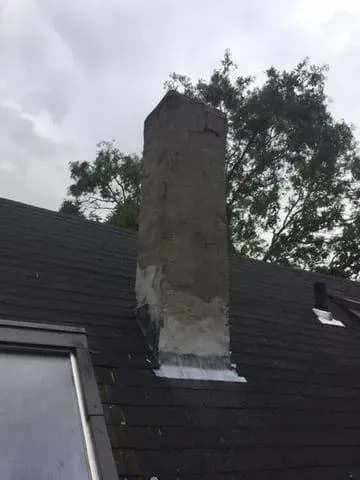 Chimney on a dark shingled roof with signs of repair, against a cloudy sky. Trees in the background suggest an outdoor setting. Calm atmosphere.