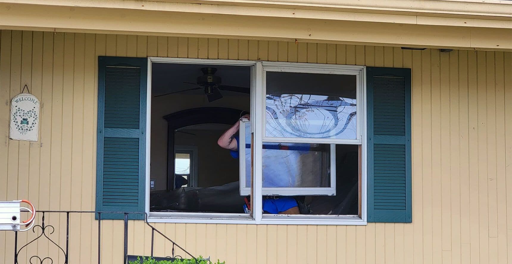 A person in a blue shirt is installing a window air conditioner from inside a beige house. Green shutters and a "Welcome" sign are visible outside.