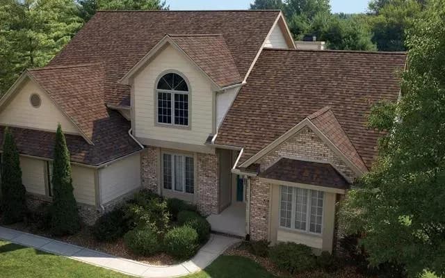 A suburban home with a steep, brown roof, light beige walls, and large windows. Surrounded by green trees and a neat lawn, conveying a peaceful ambiance.