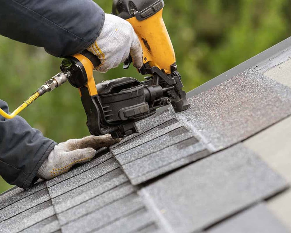 Hands in gloves using a yellow nail gun to install gray shingles on a roof. The image conveys focus and precision in construction work.