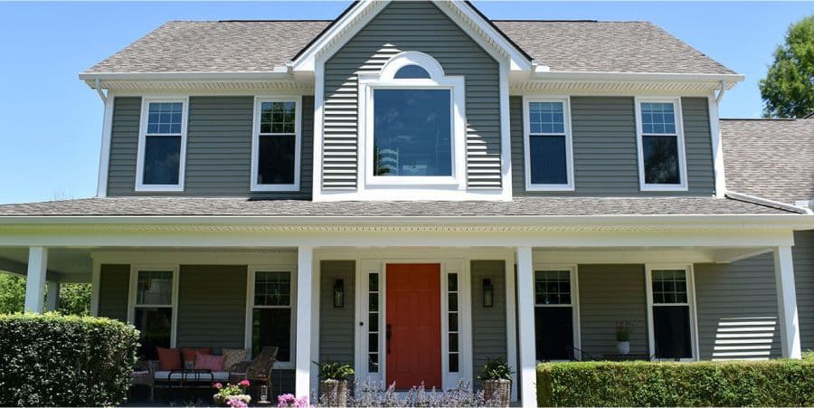 A two-story house with gray siding, white trim, and a central red door. The porch features chairs and plants. The setting is calm, under a clear blue sky.