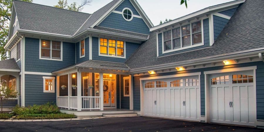 A charming two-story house with blue siding and white trim features warmly lit windows, a welcoming porch, and a spacious three-car garage at dusk.