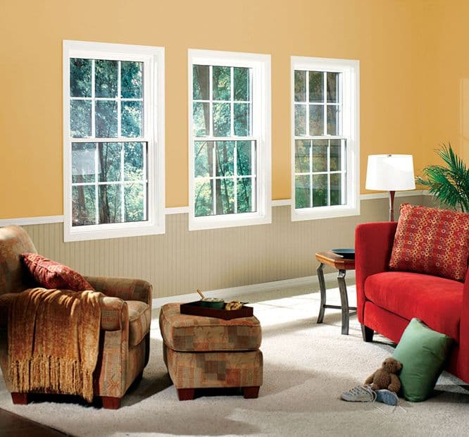 A cozy living room with three large windows showing lush greenery. Features a patterned armchair, red sofa, beige walls, and a small wooden table.