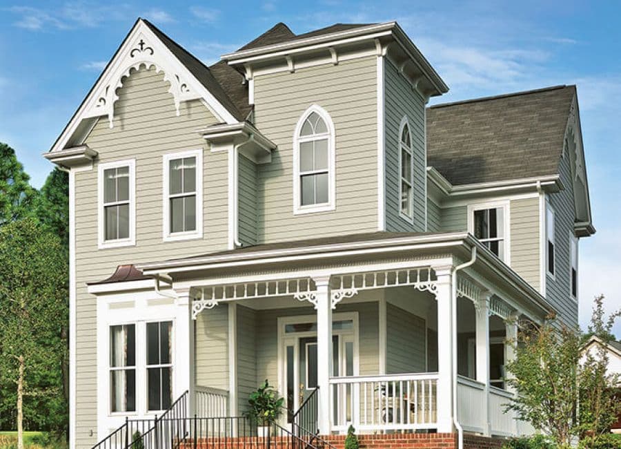 Victorian-style house with gray siding, white trim, and intricate decorative details. Features a front porch, arched window, and lush greenery.