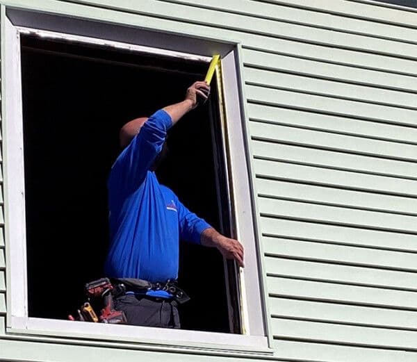 A person in a blue shirt and tool belt measures a window frame from inside a building with light green siding, using a yellow tape measure.