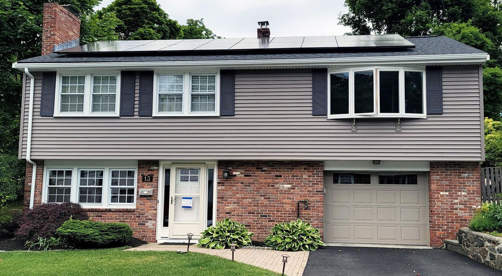 Two-story suburban house with a brick lower and siding upper facade. Solar panels on the roof, manicured lawn, and a garage to the right convey a modern, eco-friendly vibe.