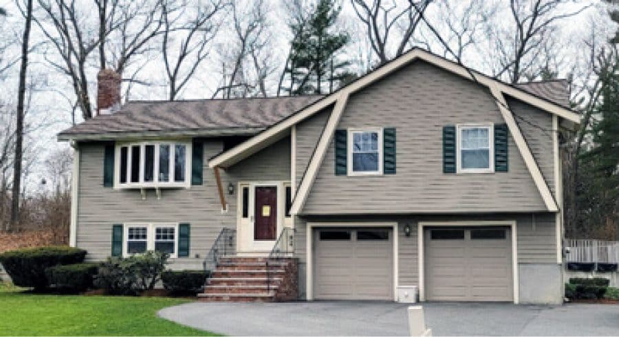 A suburban split-level house with gray siding, green shutters, and a double garage. Surrounded by bare trees and bushes on a clear day.