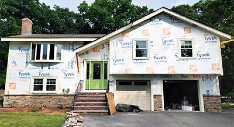 A house undergoing renovation, wrapped in Tyvek insulation. It features a prominent chimney, green doors, and an open garage. The setting is surrounded by trees.
