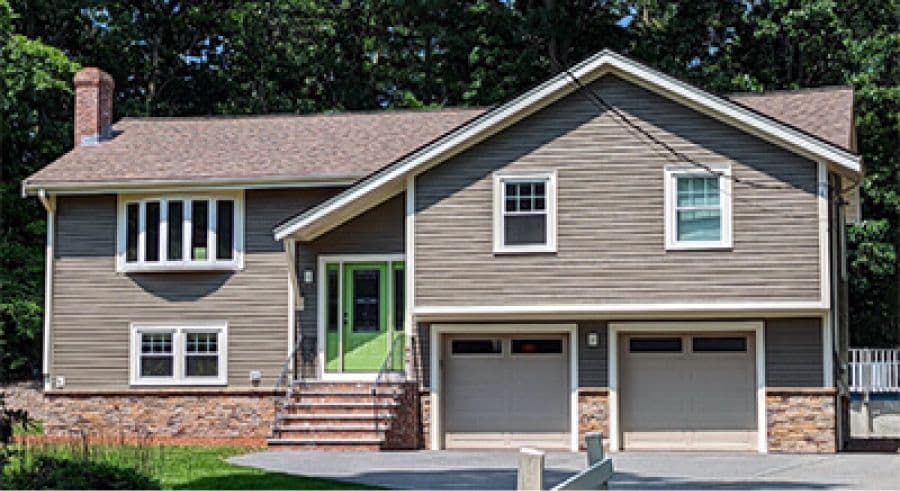 A two-story suburban house with brown siding and a green front door. It has attached double garage doors, a brick chimney, and steps leading to the entrance.