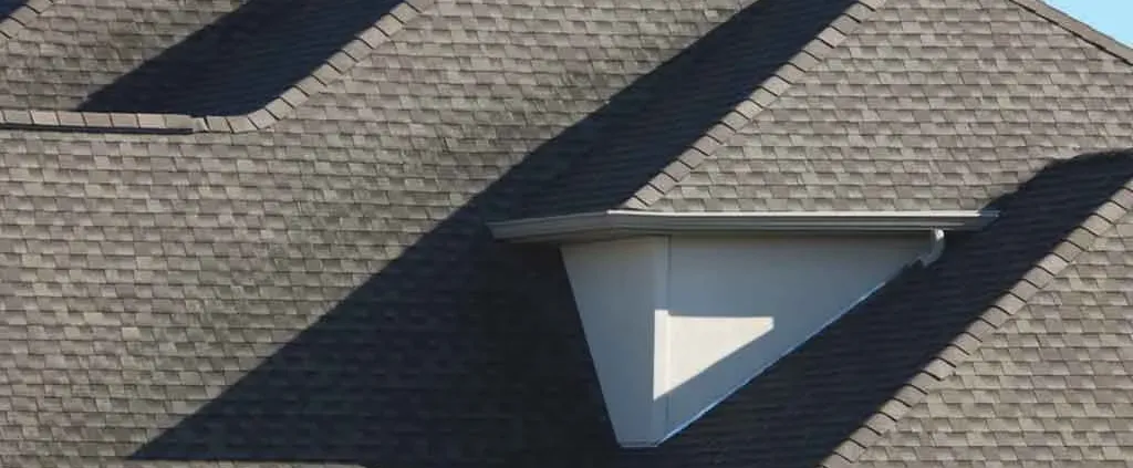 A close-up of a roof with dark gray shingles, featuring a triangular dormer window casting a shadow, creating geometric patterns.