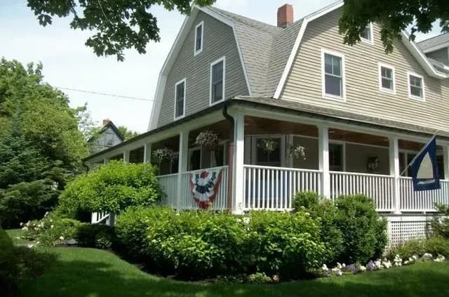 A charming, two-story house with deep green landscaping, a wraparound porch adorned with patriotic bunting, and flowering hanging baskets.