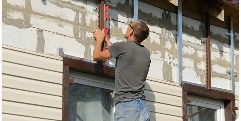 A man in a gray shirt uses a red level tool to measure the vertical alignment of siding on a house. The wall appears partially renovated.