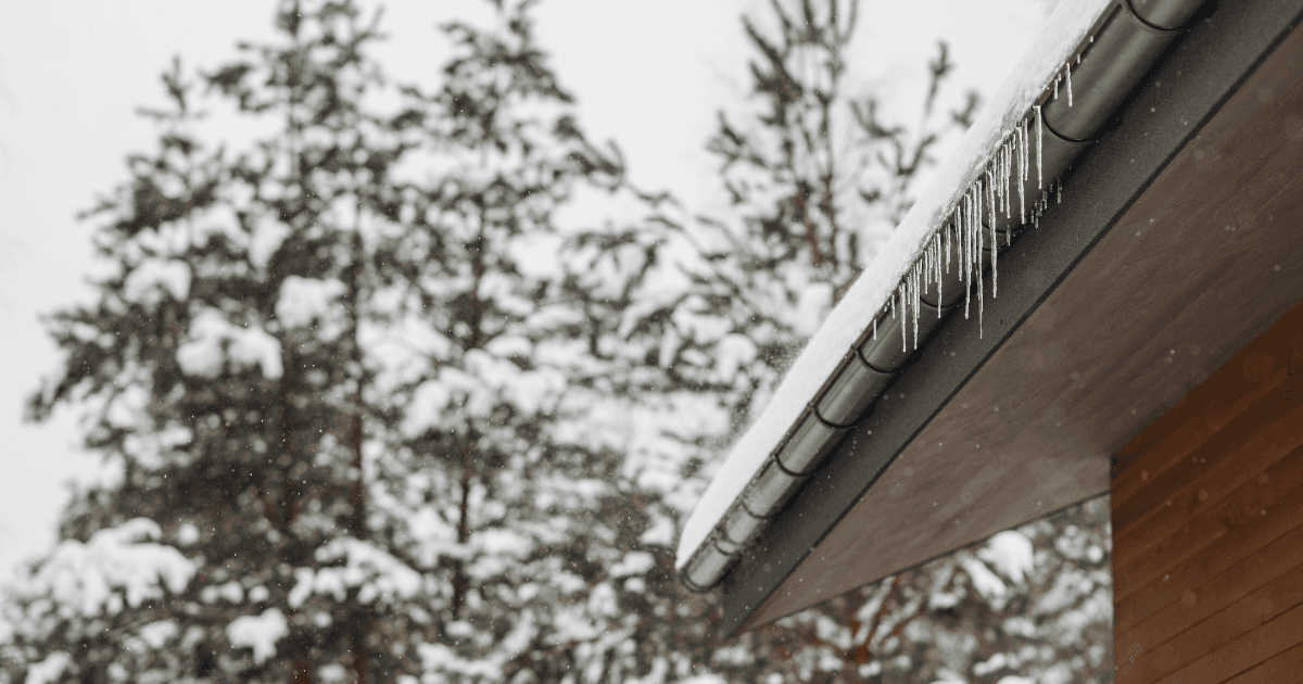 Icicles hang from the edge of a snow-covered roof with trees blurred in the snowy background, conveying a cold and serene winter scene.