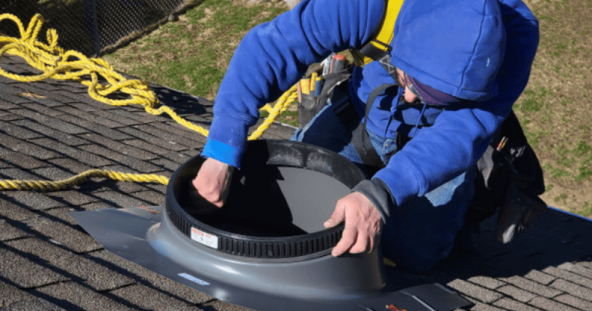 A person in a blue hoodie and safety harness installs a roof vent on shingles. They are focused, with tools nearby and ropes for safety.