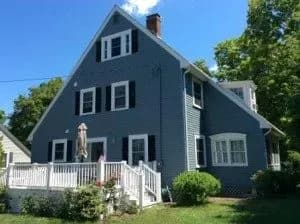 A blue, two-story A-frame house with white trim and a front porch, surrounded by lush greenery, under a clear blue sky.