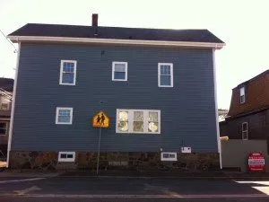 A blue house with small windows and missing doors faces a street. A leaning pedestrian crossing sign is in front, creating a surreal, quirky scene.