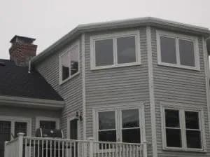 A two-story house with gray siding and large windows. The second floor has floor-to-ceiling windows, and a chimney is visible on the roof. The overcast sky gives a serene mood.