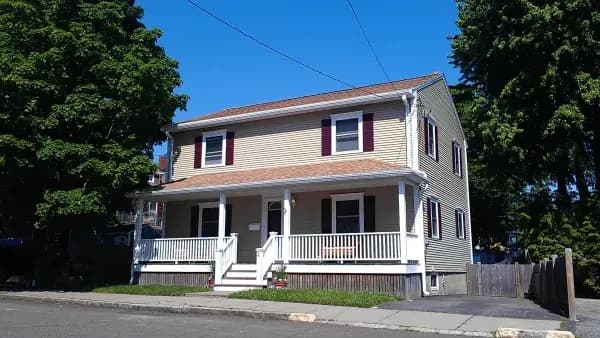 Two-story beige house with a red roof and shutters, large porch, and lush trees on a clear day. White railing and steps lead to the entrance.