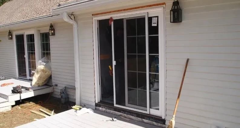 A white sliding glass door is being installed on a house with beige siding. Tools and insulation are nearby. The scene conveys home renovation.