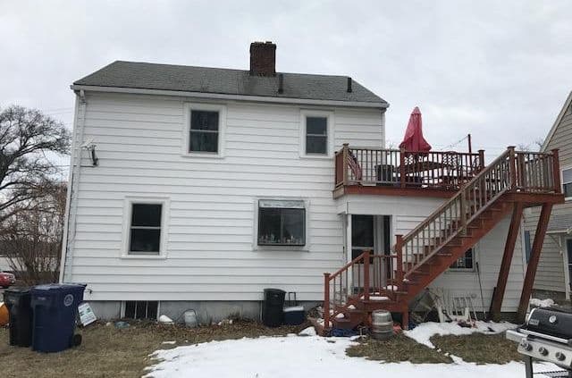 Two-story white house with snowy yard, outdoor stairs leading to a second-floor deck with a red umbrella. Overcast sky, bare trees, relaxed vibe.