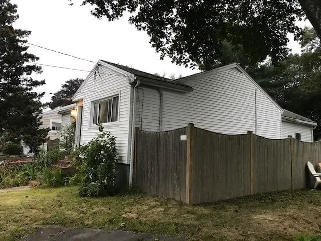 Single-story white house with a sloped roof, partially obscured by a wooden fence. Surrounding greenery and overcast sky create a calm atmosphere.