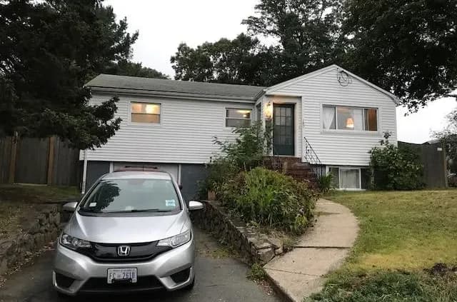 A white single-story house with a grey roof and a front yard featuring a path and shrubs. A silver Honda car is parked in the driveway. Overcast sky.