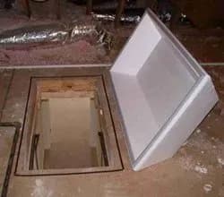 Open trapdoor in a wooden floor reveals a staircase leading down. A white foam cover rests beside the entrance. The setting is dim and dusty.