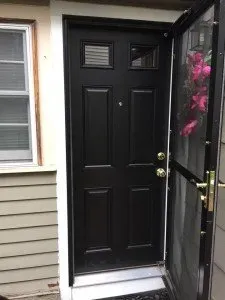 A black six-panel door with brass handles, partially open, revealing a glass storm door. Pink flowers are visible through the glass. Adjacent is a window.