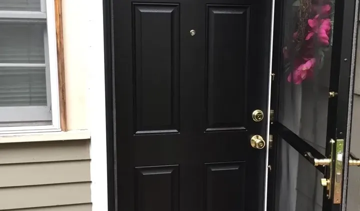 A closed black door with decorative paneling and a shiny brass handle, set within a beige exterior wall. A wreath with pink flowers hangs on a storm door.