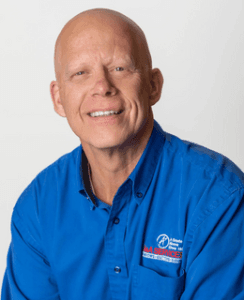 A smiling person in a bright blue shirt with company logos sits against a plain white background, conveying a friendly and approachable demeanor.