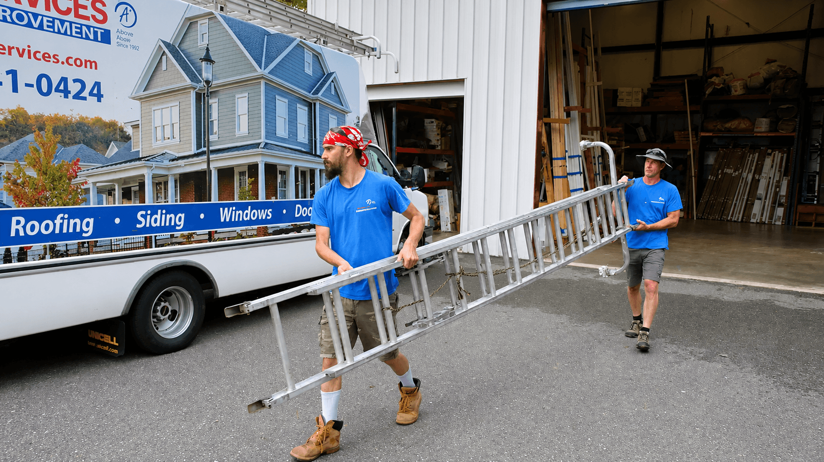 Two workers in blue shirts carry a ladder from a truck displaying home improvement services. The scene conveys teamwork and outdoor labor.
